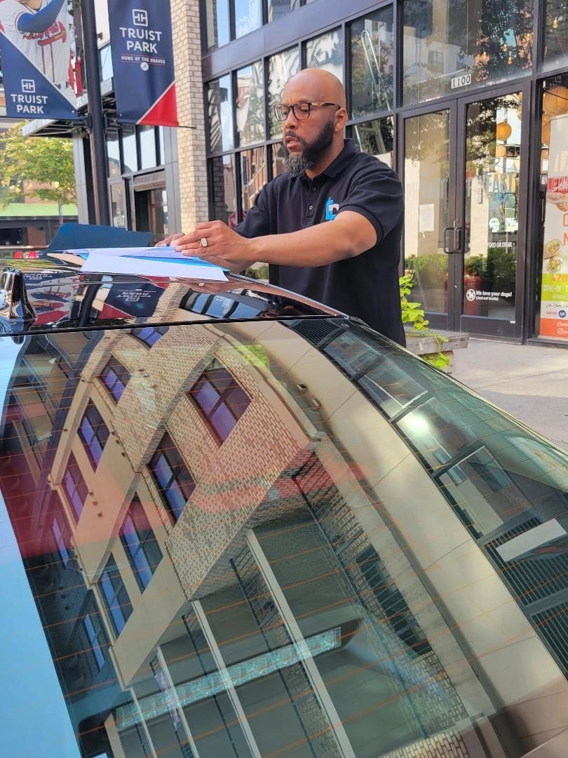 William signing papers at car hood on urban street